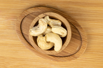 Lot of whole brown nut cashew in bamboo bowl on round bamboo coaster flatlay on light wood