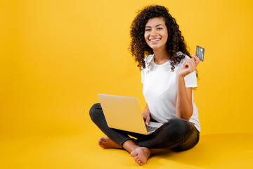 happy african black woman sitting with laptop and credit card isolated over yellow