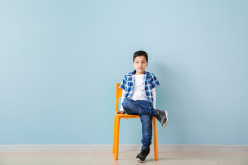 Cute fashionable boy sitting on chair against color wall
