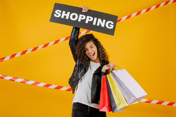 black woman with shopping sign and colorful shopping bags isolated over yellow with signal tape