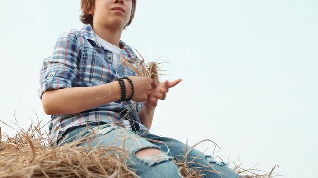 Teenager boy in jeans and checkered shirt sitting on haystack on countryside field. Portrait handsome boy on rural haystack on harvesting field in village