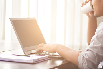 Selective focus of the hand of business man using laptop and drinking coffee at office building with warm light. The asian man are working notebook on the wooden table.