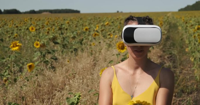 Girl alone in whole field with sunflowers has fun wearing virtual reality glasses, sniffs sunflower, throws it, runs around field, plays, enjoys virtual vision, helmet on her head, beautiful landscape