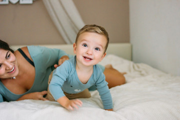 Charming happy little baby boy having fun with mom brunette woman on bed in bright bedroom