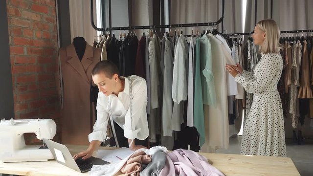 caucasian tailor work on new womenswear collection for client in cozy workshop studio. Client in white dress stands near clothing rack with fashionable stylish clothes made by fashion designer.
