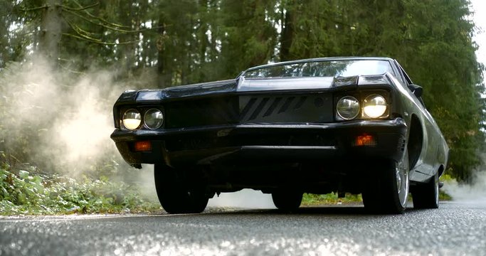Front view daring look of a black classic sports American car on the road in the woods is wound up and fangs of exhaust gases