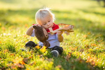 Beautiful blonde two years old toddler boy, playing with wooden plane and teddy bear in the park on sunset