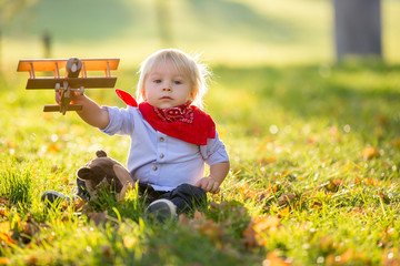 Beautiful blonde two years old toddler boy, playing with wooden plane and teddy bear in the park on sunset