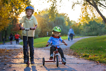 Beautiful blonde two years old toddler boy and his older brother, riding red tricycle and scooter in the park on sunset