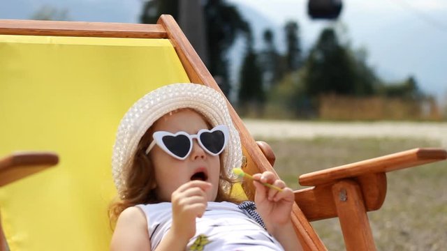 Tourist baby girl is sitting on a deck chair. Baby girl with sunglasses and a hat eats cand y and relaxes on a journey