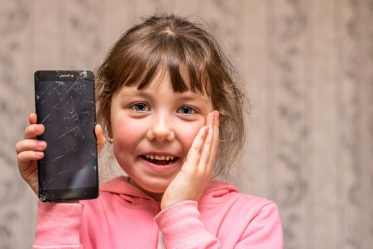  The child broke the telephone communicator and laughs. Baby joked breaking smartphone screen. Portrait of a cheerful little girl with a mobile phone.