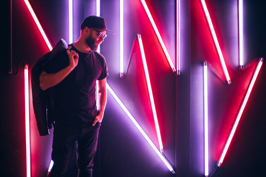 Hipster handsome man on the city streets being illuminated by neon signs. He is wearing leather biker jacket or asymmetric zip jacket with black cap, jeans and sunglasses.