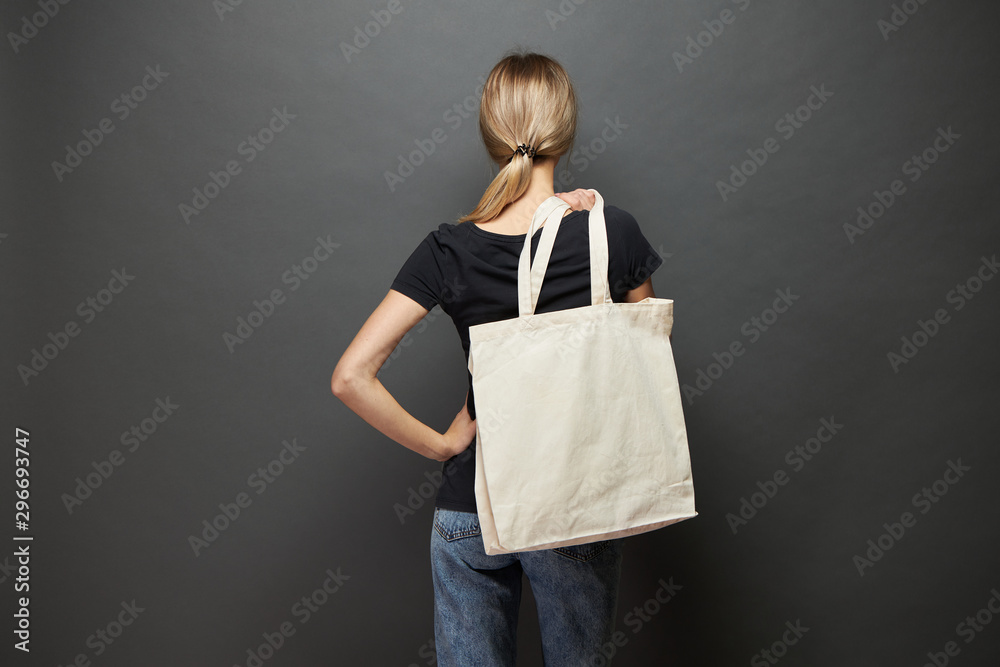 Wall mural Young woman holding white textile eco bag against gray background. Ecology or environment protection concept. White eco bag for mock up.