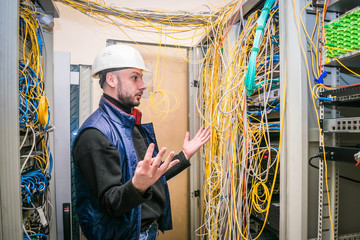 Surprised engineer in white helmet spreads his arms to the side near the rack with server...
