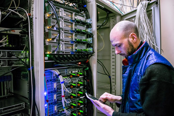 A man with a digital tablet working in a server room. The engineer stands next to a multitude of...