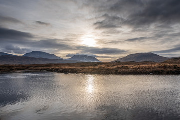 Wetlands and wintery grass fields as the sun starts to set over the Black Mount mountain range on a partially cloudy day in the Scottish Highlands.