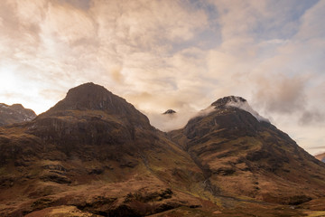 Two of the three mountain peaks called The Three Sisters, covered in brown wintery vegetation on a partially cloudy day in Scottish Highlands.