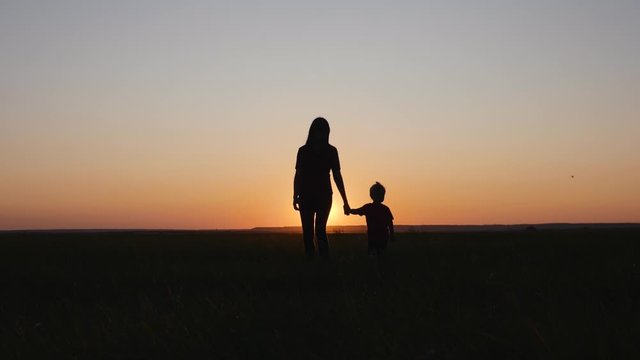 Silhouette of a mother and child walking across a field at sunset. Happy family outdoors in summer, walking on their day off.