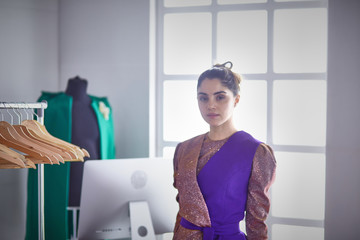 Beautiful young stylist holding clipboard near rack with designer clothes on white background