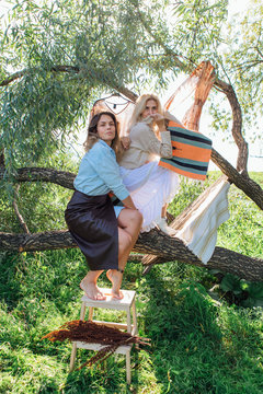 Portrait Of Two Beautiful Women Brunette And Blond Sitting On The Big Brunch Of Tree In A Field In Late Summer. Beautiful Girls Standing Next To Each Other.