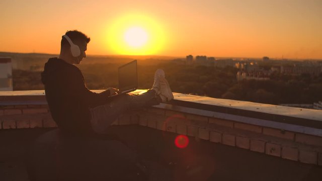 A programmer with a laptop in headphones sits on the roof and writes code at sunset and listens to music. Remote work freelancer. Freedom to work, Typing on keyboard at sunset with a view of the city