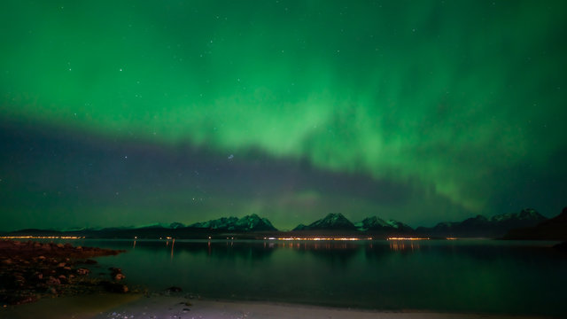 Green northern ligths almost covering the sky above mountain range. Beach in foreground. Many stars. Aurora, mountains and city lights reflected in water. Tromso, Norway.