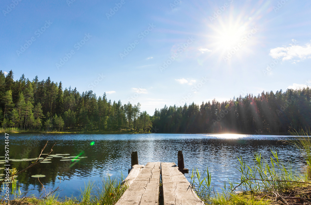 Wall mural Traditional Finnish and Scandinavian view. Beautiful lake on a summer day and an old rustic wooden dock or pier in Finland. Sun shining on forest and woods in blue sky.