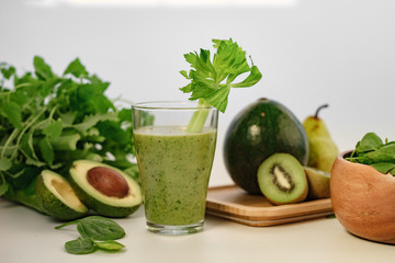 Green smoothie with a stalk of celery in a glass cup. On the table is an avocado.