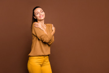 Photo of cheerful cute friendly lovely sweet pretty cute youngster hugging herself smiling toothily beaming enjoying her newly bought warm sweater wearing trousers isolated brown pastel color
