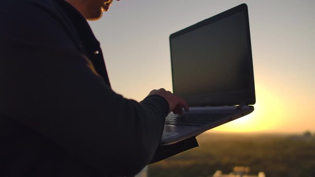 A programmer with a laptop sits on the roof and writes code at sunset. Remote work freelancer. Freedom to work. Typing on a keyboard at sunset with a view of the city