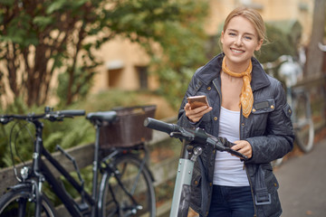 Happy young woman in casual grey jacket looking at camera and smiling, standing in the street with an e-scooter with mobile phone in her hands.