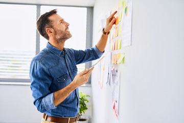 Web developer looking at notes on office wall and holding tablet