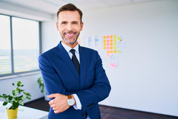 Confident businessman smiling at camera with arms crossed while standing in office - obrazy, fototapety, plakaty