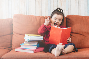 Thoughtful little asian girl reading a book on a sofa. Concept of child growth, education and...