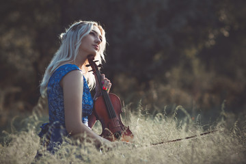 portrait of a young fabulously beautiful girl in a dress with a violin sitting in dry grass on meadoe at the dawn, woman playing a musical instrument with inspiration relaxing on nature © fantom_rd