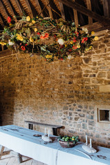 Une salle de réception dans un château médiéval. Une table décorée dans un château du Moyen-Age. Une couronne de fleurs suspendue au dessus d'une table de repas. Une salle à manger dans un château.