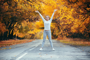slim young woman in sportswear exercises and stretches hands up satanding on asphalt road, girl engaged in sport outdoors on background of autumn foliage, concept healthy lifestyle and female beauty