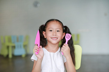 Happy little Asian child girl showing plastic knife and spade of frying pan in play room ready to cook learning.