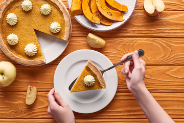 cropped view of woman holding spatula with piece of pumpkin pie with whipped cream near baked pumpkin, cut and whole apples on orange wooden table