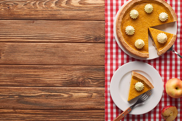 delicious pumpkin pie with whipped cream on checkered tablecloth near fork, cut and whole apples on brown wooden table
