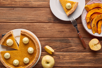 cut pumpkin pie with whipped cream near baked pumpkin, whole and cut apples and fork on brown wooden table