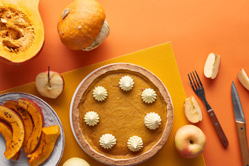 tasty pumpkin pie with whipped cream near raw and baked pumpkins, whole and cut apples, knife and fork on orange surface