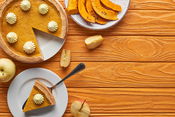 delicious pumpkin pie with whipped cream near baked pumpkin, whole and cut apples on orange wooden table
