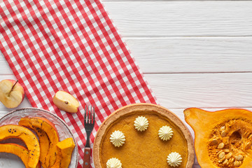 tasty pumpkin pie with whipped cream on checkered napkin near baked and raw pumpkins, cut apple and fork on white wooden table