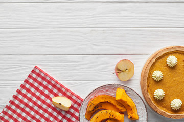 tasty pumpkin pie with whipped cream near baked sliced pumpkin, cut apple and checkered napkin on white wooden surface