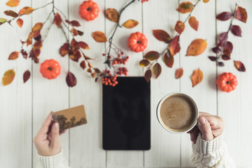 Naklejka premium Woman with tablet and credit (discount) card cup on white retro wood boards. Coffee, tablet, leaves, rowan and small pumpkins . Autumn, fall concept. Flat lay, top view.