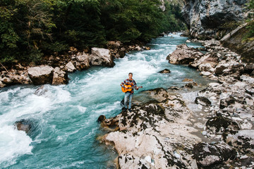 Man playing guitar standing on the bank of a mountain river on a background of rocks and forest. Handsome hippie style guitarist engrossed on music outdoors. Concept of freedom relaxation. Place for