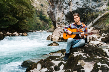 Young man playing guitar sitting on the bank of a mountain river on a background of rocks and...