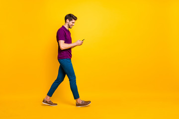 Full length body size profile side view of his he nice attractive cheerful cheery glad addicted guy using digital device 5g smm walking isolated over bright vivid shine vibrant yellow color background