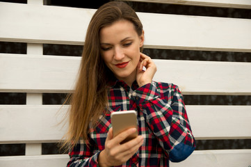 Young beautiful girl stands near a white wall, smiles and holds a mobile phone, looks at the phone screen.